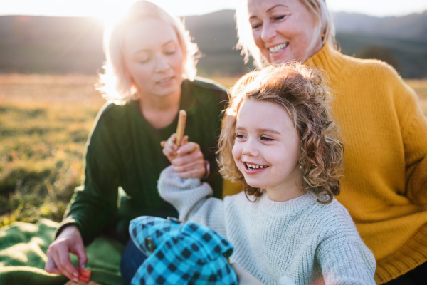 Happy small girl with mother and grandmother having picnic in nature at sunset.