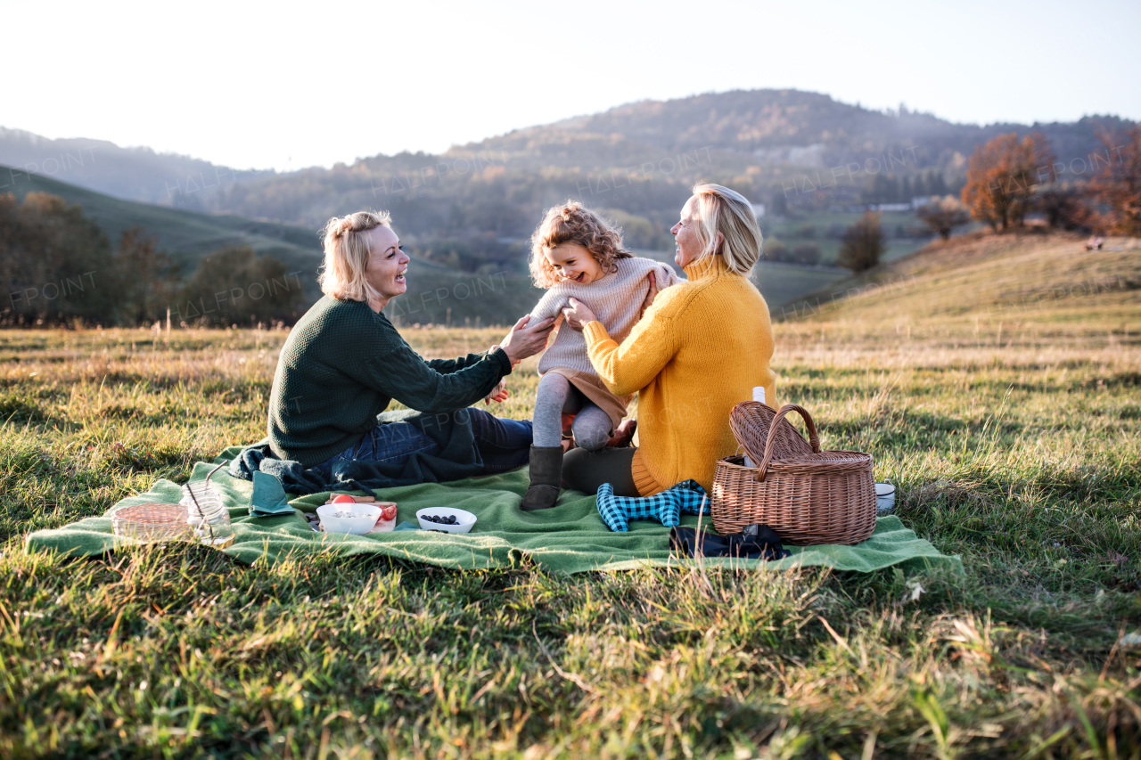 Happy small girl with mother and grandmother having picnic in nature at sunset.