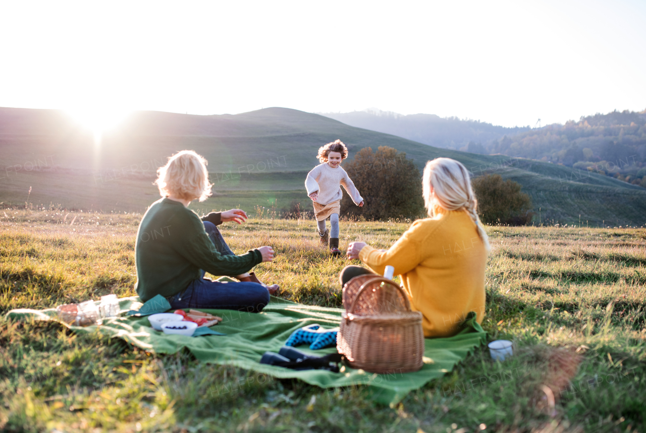 Happy small girl with mother and grandmother having picnic in nature at sunset.