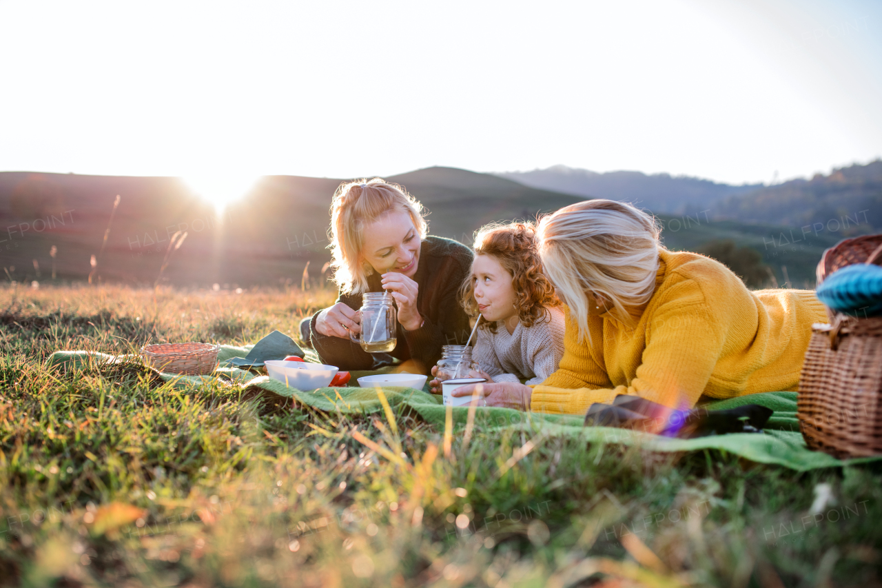 Happy small girl with mother and grandmother having picnic in nature at sunset.