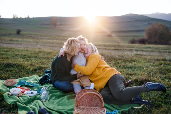 Happy small girl with mother and grandmother having picnic in nature at sunset, having fun.