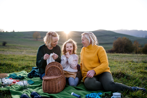 Happy small girl with mother and grandmother having picnic in nature at sunset.