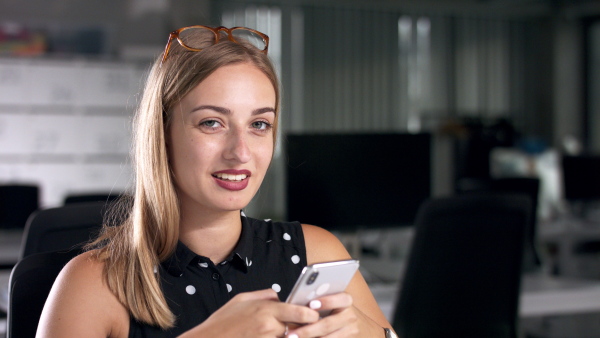 Young businesswoman with smartphone indoors in office, looking at camera. Coronavirus concept.