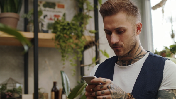 Man florist standing in flower and plant shop, using smartphone.