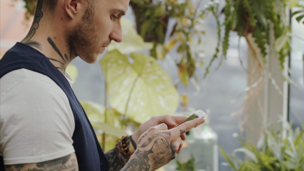 Young man florist standing in flower and plant shop, using smartphone.