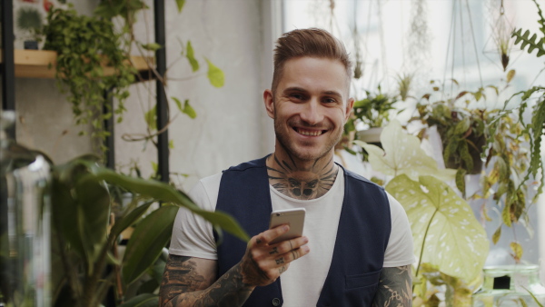 Man florist with smartphone standing in flower and plant shop, looking at camera.