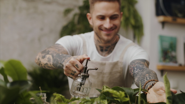 Young man florist standing in flower and plant shop, spraying plants with water.