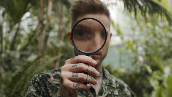 Young man researcher with magnifying glass standing in botanical garden, looking at camera.