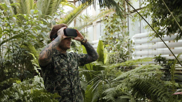 A young man standing in botanical garden, using VR glasses.