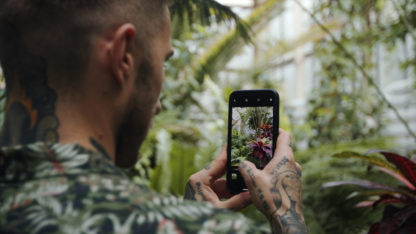 Midsection of young man standing in botanical garden, taking photographs with smartphone.