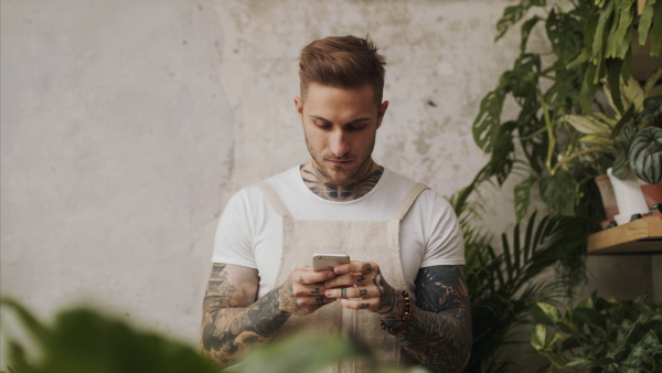 Young man florist standing in flower and plant shop, using smartphone.