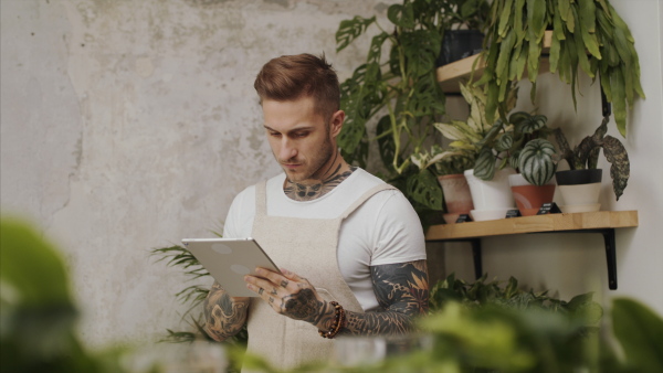 Young man florist standing in flower and plant shop, using tablet.