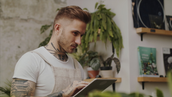 Young man florist standing in flower and plant shop, using tablet.