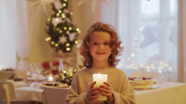 Front view of cheerful small girl standing indoors at Christmas, holding candle.