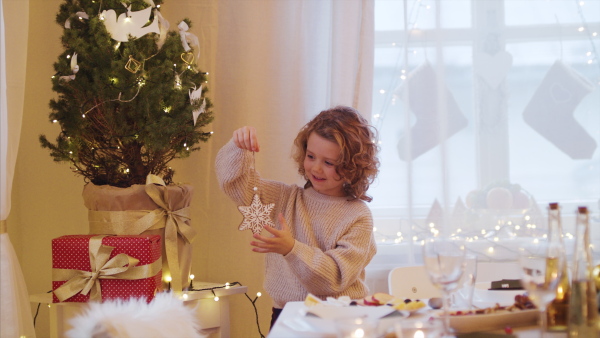 Front view of happy small girl indoors at home, standing by Christmas tree.