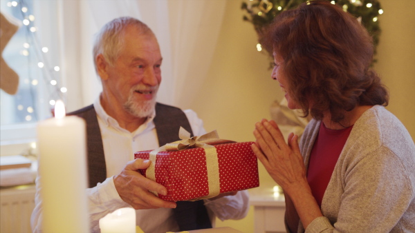 Portrait of happy senior couple in love indoors at home sitting at the table at Christmas, giving gifts.