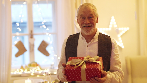 A front view of happy senior man indoors holding present at Christmas.