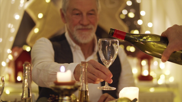 Senior man with family sitting at the table indoors at Christmas, dinner and wine pouring concept.