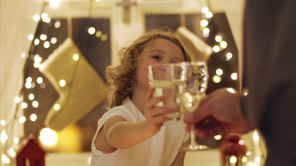 Happy small girl with family sitting indoors celebrating Christmas together, clinking glasses.