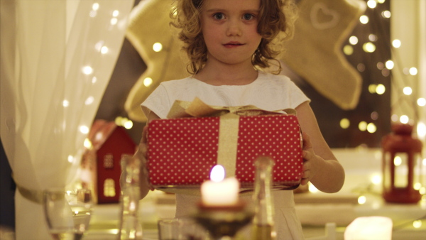 Front view of cheerful small girl indoors at the table at Christmas, holding present.