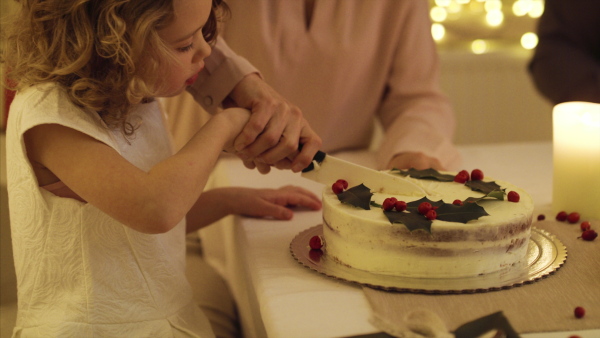 Small girl with unrecognizable parents at the table indoors celebrating Christmas together, cutting cake.