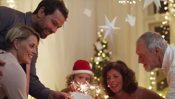 Happy small girl with parents and grandparents indoors celebrating Christmas, holding sparklers.