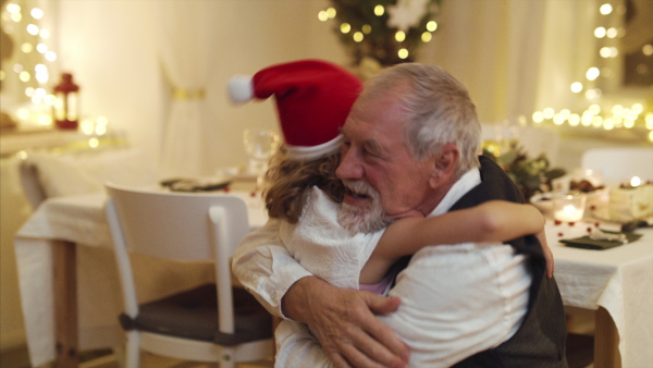 Cheerful small girl with senior grandfather indoors at home at Christmas, hugging.