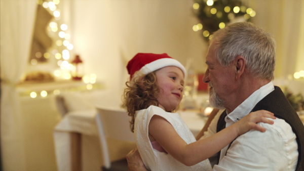 Cheerful small girl with senior grandfather indoors at home at Christmas, hugging.