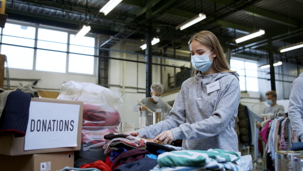 Portrait of volunteers sorting out donated clothes in community charity donation center, looking at camera. Coronavirus concept.