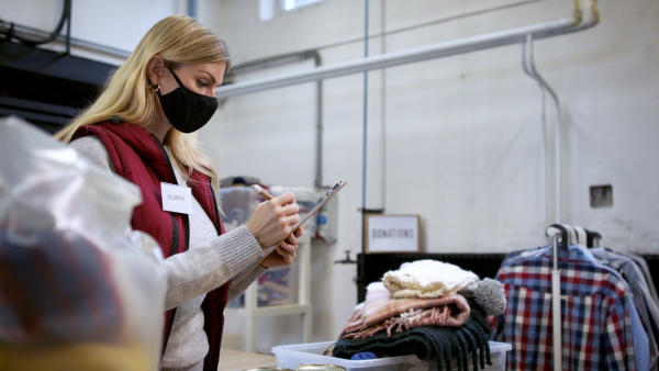 Woman volunteer working with clothes in community charity donations center, looking at camera. Coronavirus concept.