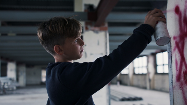 Side view of teenager boy indoors in abandoned building, using spray paint on wall.