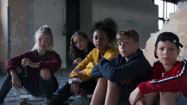 Front view of group of teenagers gang sitting indoors in abandoned building, looking at camera.