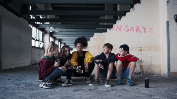 Front view of group of teenagers gang with smartphone talking indoors in abandoned building, looking at camera.