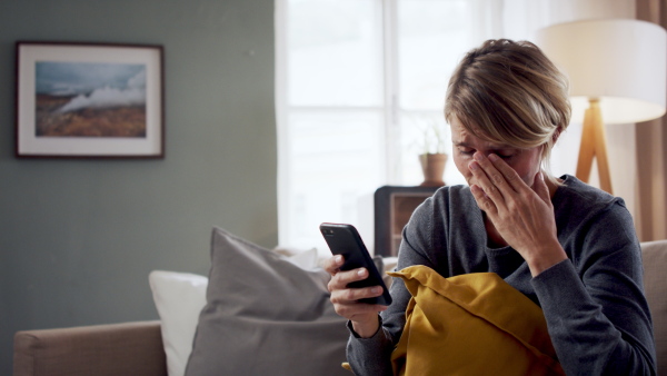 Worried woman with smartphone indoors on sofa at home feeling stressed, mental health concept.