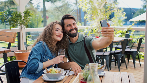 Happy couple sitting outdoors on terrace restaurant, taking selfie with smartphone.