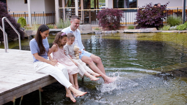 Cheerful family with children sitting outside hotel on holiday, bare feet in water in lake.