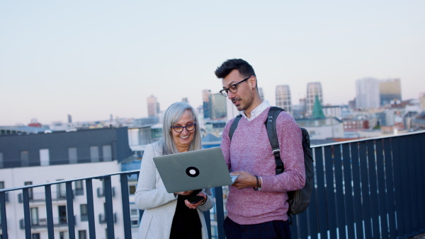 Young and old businesspeople working outdoors on terrace in city, using laptop.