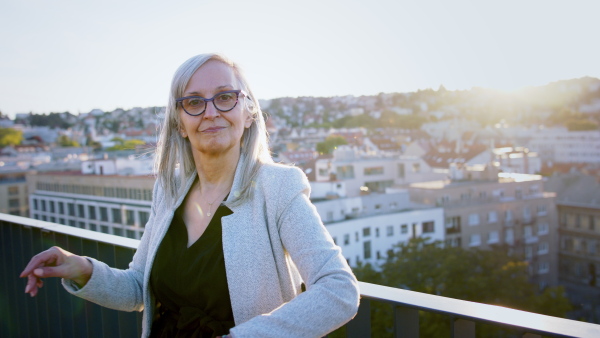 Senior businesswoman standing outdoors on building terrace, leaning on railing.