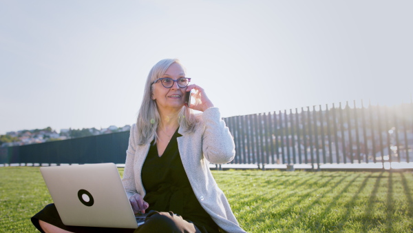 Old businesswoman working outdoors in city, sitting on grass and using laptop and smartphone.