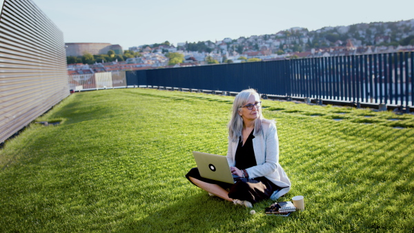 Old businesswoman working outdoors in city, sitting on grass and using laptop.