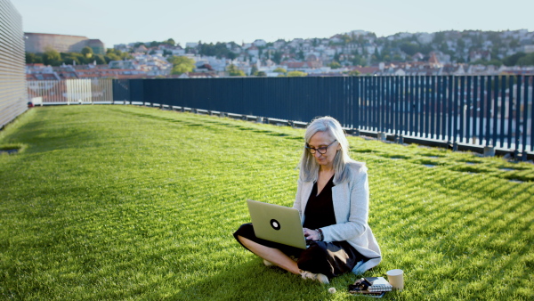 Old businesswoman working outdoors in city, sitting on grass and using laptop.