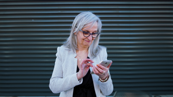 Portrait of senior businesswoman standing outdoors against dark background, using smartphone.