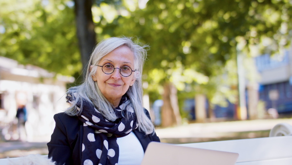 Senior businesswoman sitting and working outdoors on bench in city, using laptop and looking at camera.