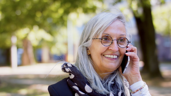 Portrait of senior woman sitting and working outdoors on bench in city, using smartphone.