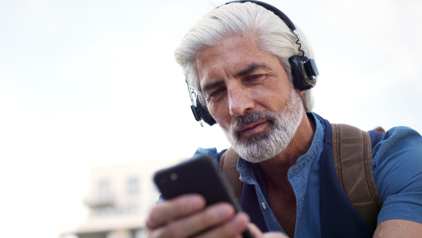 Close-up portrait of mature man with headphones sitting outdoors in city, using smartphone.