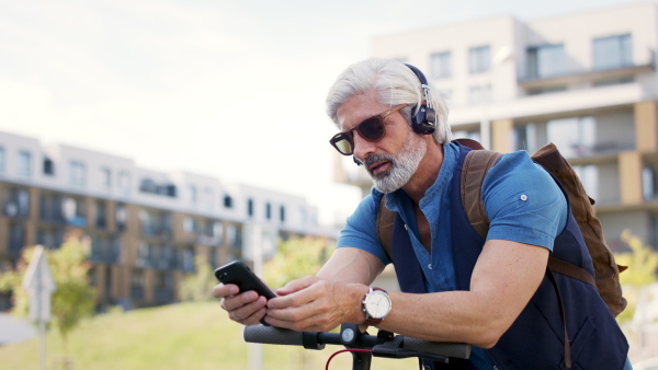 Portrait of mature man commuter with electric scooter outdoors in city, using smartphone and headphones.