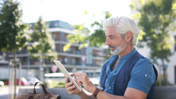 Side view portrait of mature man sitting outdoors in city, using tablet.