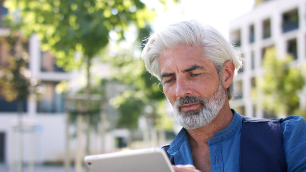 Front view portrait of mature man sitting outdoors in city, using tablet.