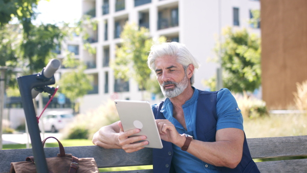 Portrait of mature man with electric scooter sitting on bench outdoors in city, using tablet.