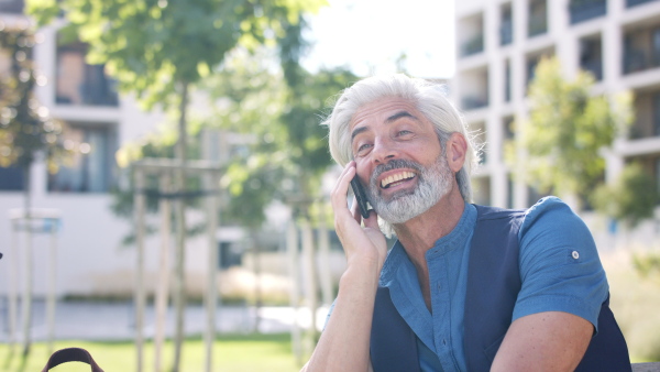 Portrait of cheerful mature man with smartphone sitting outdoors in city, making a phone call.